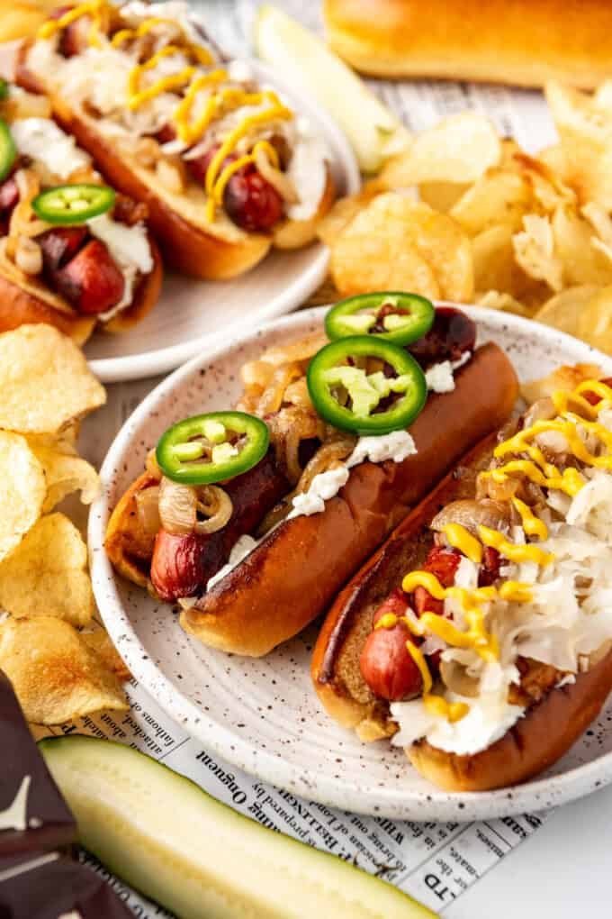 Hot dogs on buns with cream cheese, sliced jalapenos, and caramelized onions  with chips and pickle spears surrounding them.