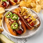 An image of Seattle-style hot dogs on a plate with a pickle spear in front and chips in the background.