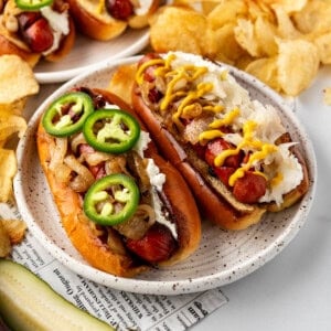 An image of Seattle-style hot dogs on a plate with a pickle spear in front and chips in the background.
