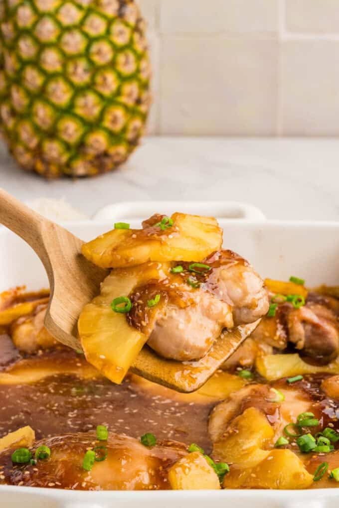 A serving spoon lifting chicken thighs and pineapple from a baking dish.