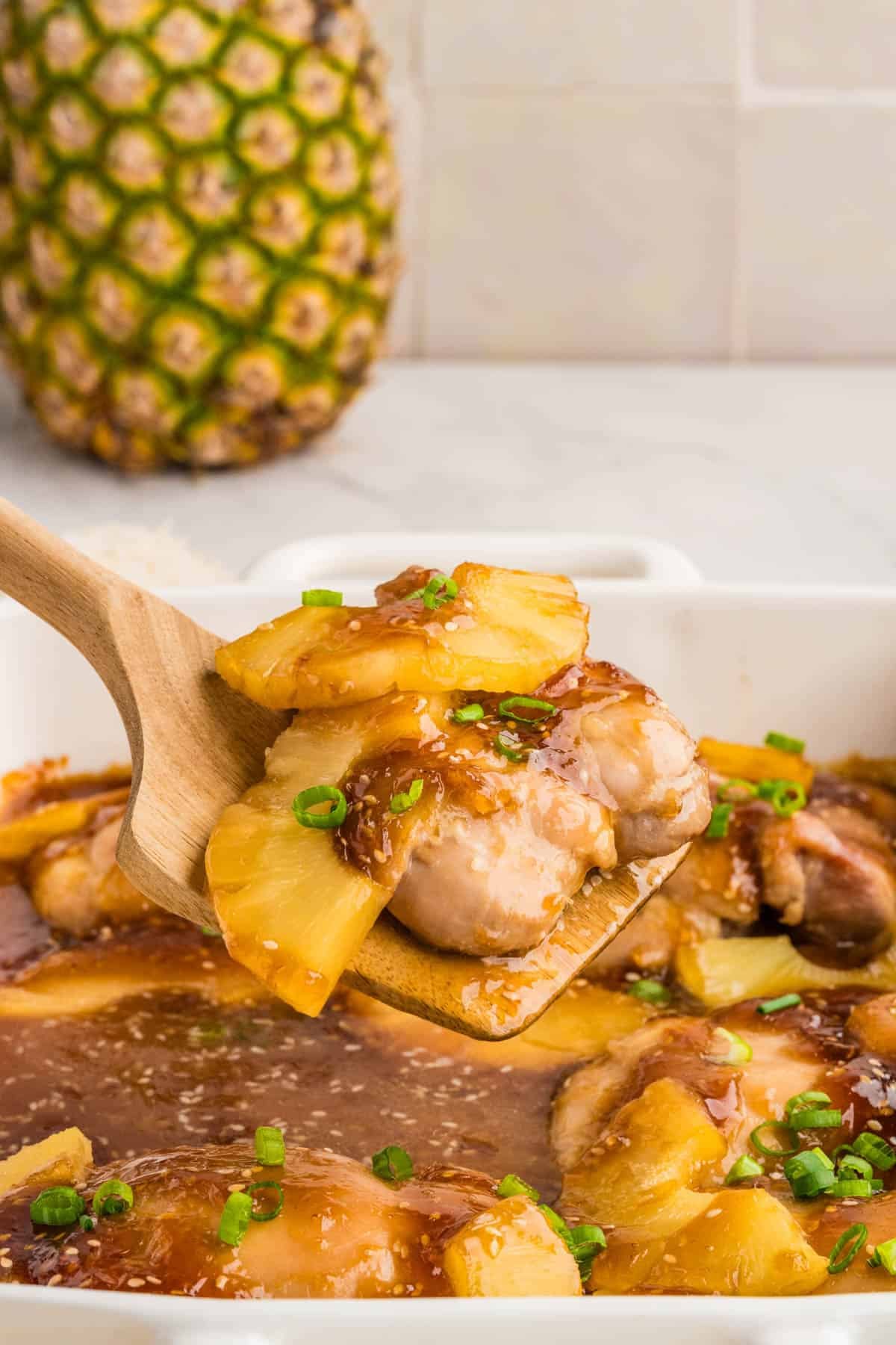 A serving spoon lifting chicken thighs and pineapple from a baking dish.