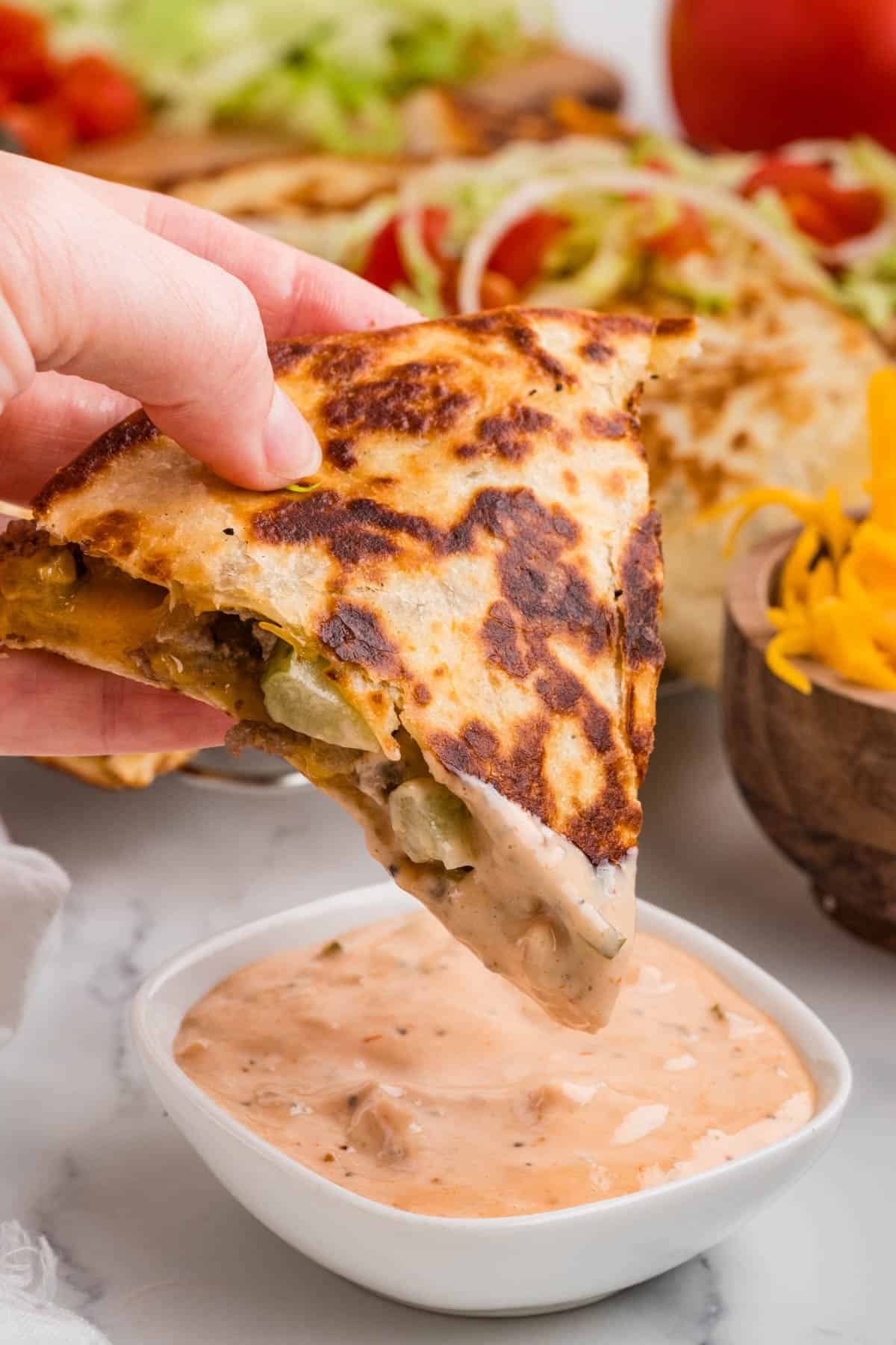 A hand holding a triangle of quesadilla over a bowl of burger sauce for dipping.