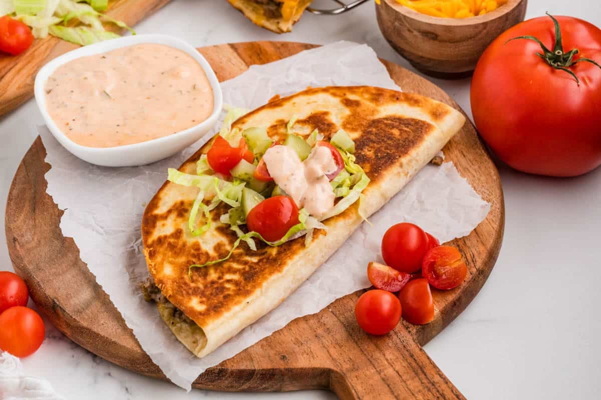 A cheeseburger quesadilla on a wooden cutting board with burger sauce in the background and tomatoes in front.