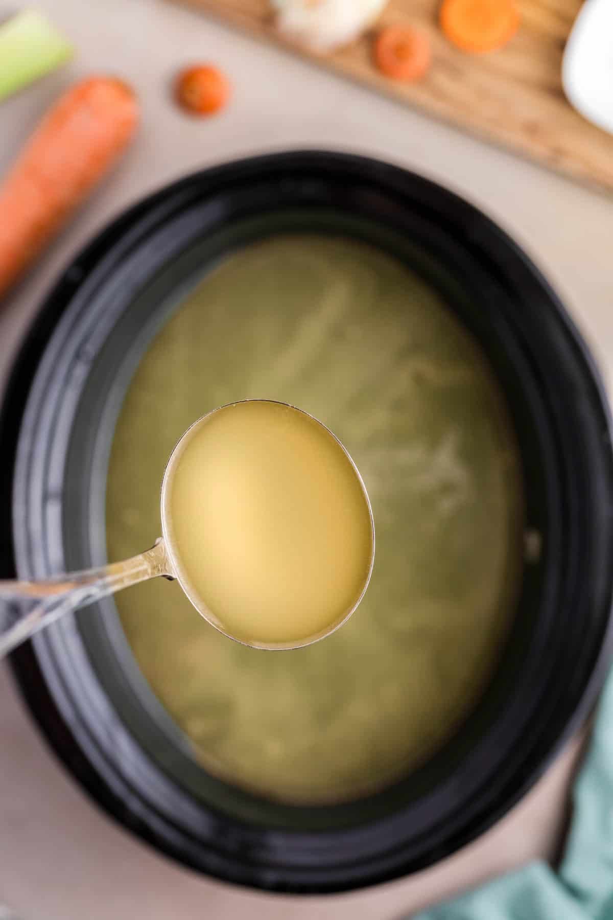 An overhead image of a ladleful of homemade chicken broth being lifted over a slow cooker.