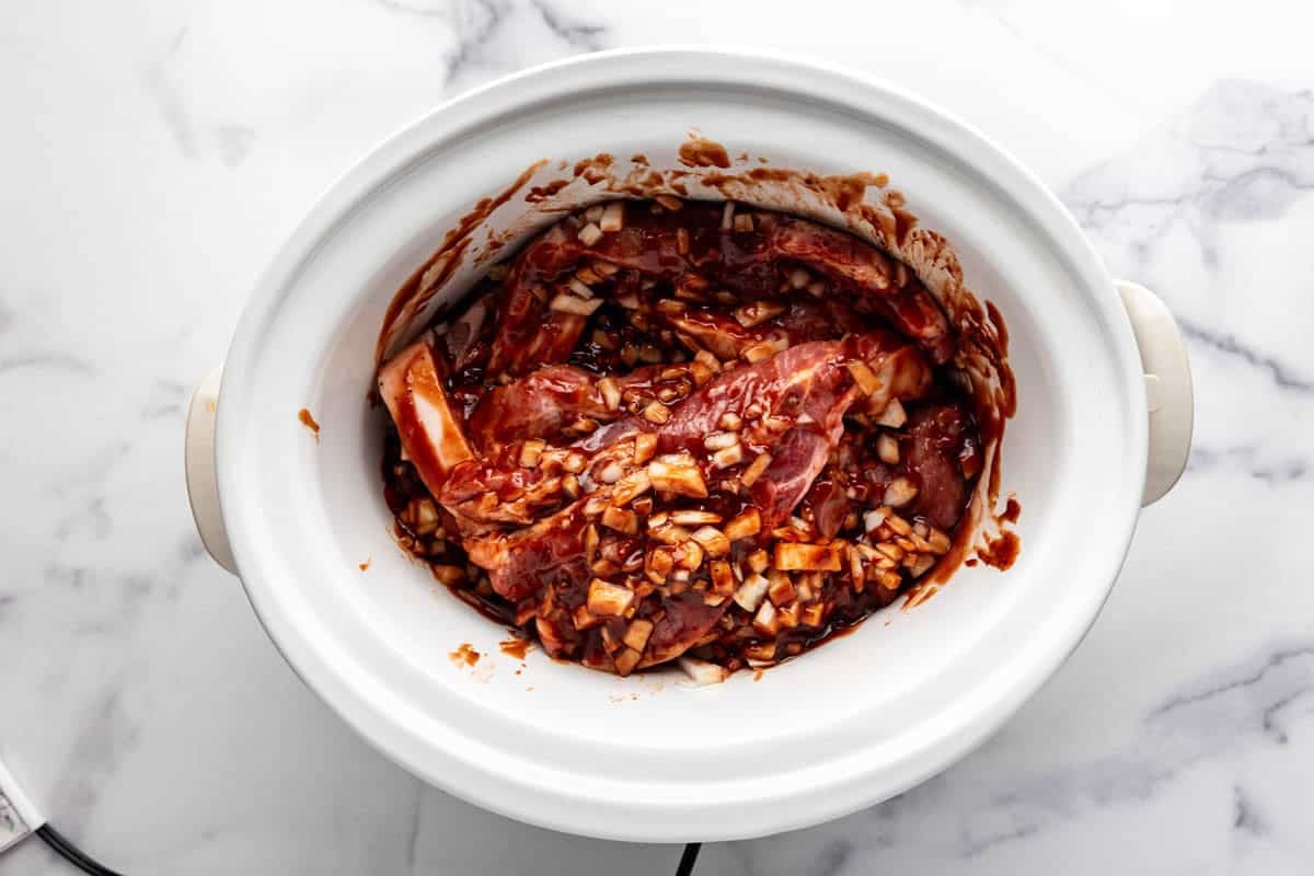 Combining country style ribs in a white crockpot with bbq sauce, brown sugar, onions, and other ingredients.