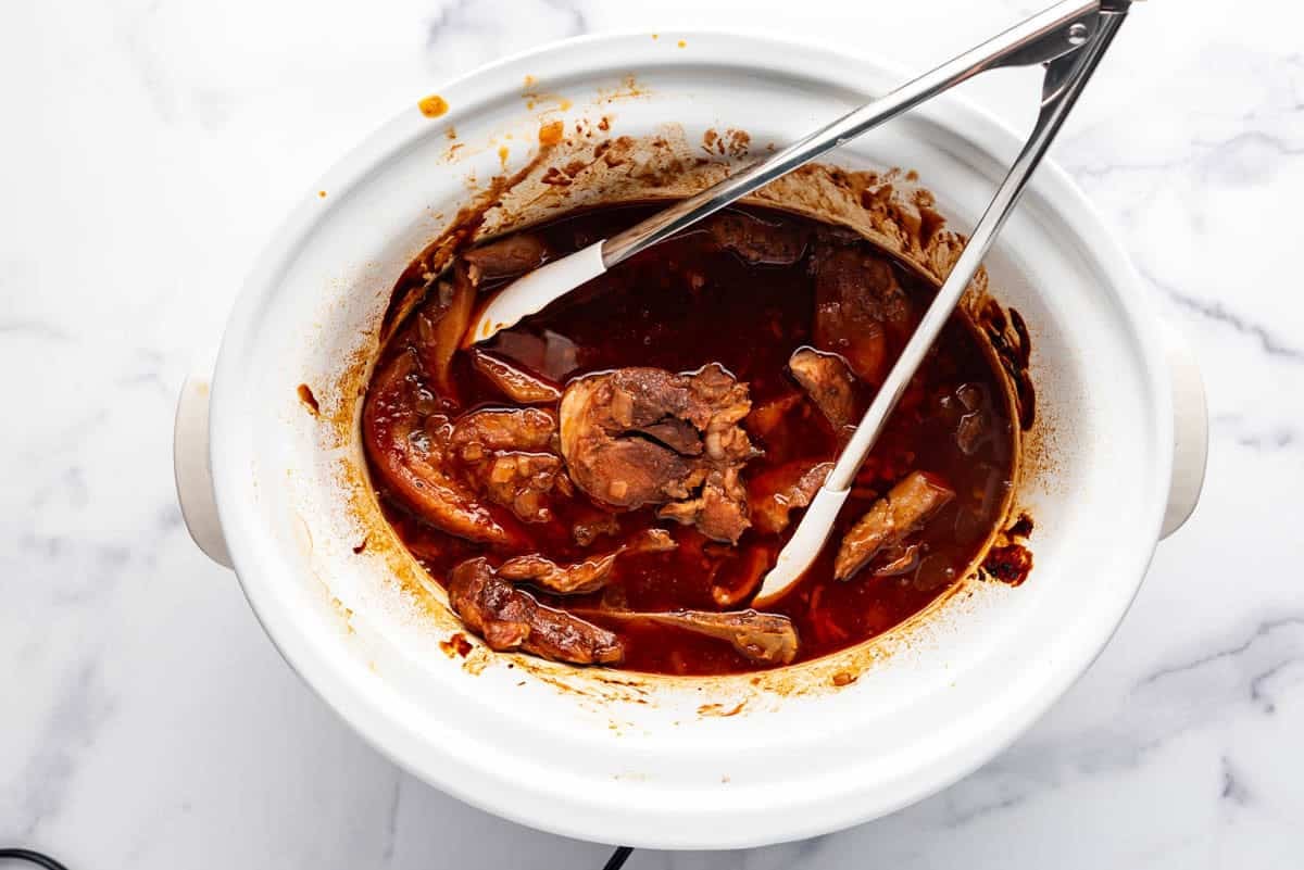 A slow cooker full of country style ribs in bbq sauce with tongs in it.