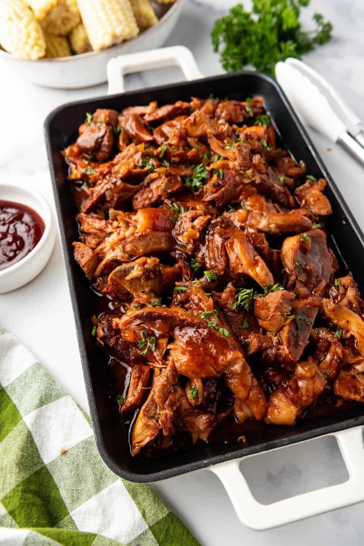 Tender country style ribs on a platter next to a bowl of bbq sauce with corn on the cob in the background.