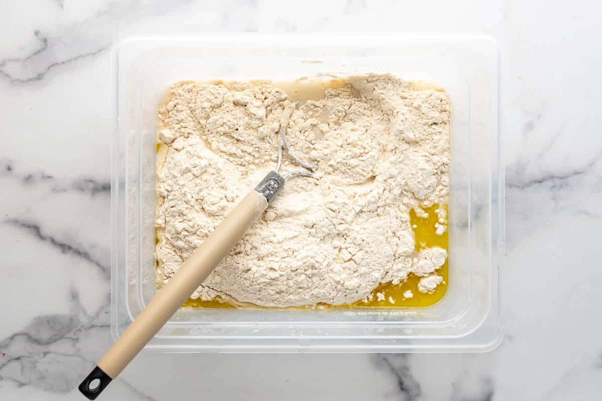 Combining water, sourdough starter, bread flour, oil, and salt in a large container with a bread whisk.