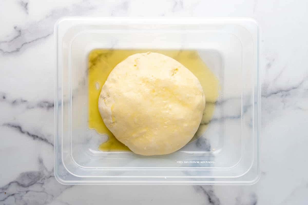 A smooth ball of sourdough pizza dough in a container with olive oil drizzled on top of the dough.