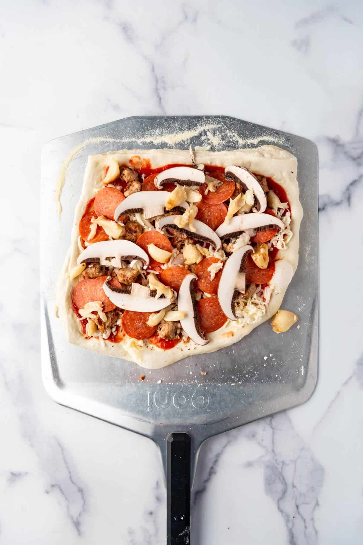 An overhead image of sourdough pizza dough topped with pepperoni, sausage, sliced portabello mushrooms, roasted garlic, and cheese on a pizza peel ready to go into the pizza oven to be baked.