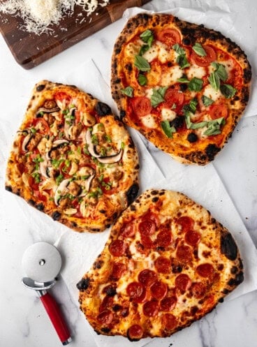 An overhead image of three different sourdough pizzas on a white surface with a pizza cutter nearby.